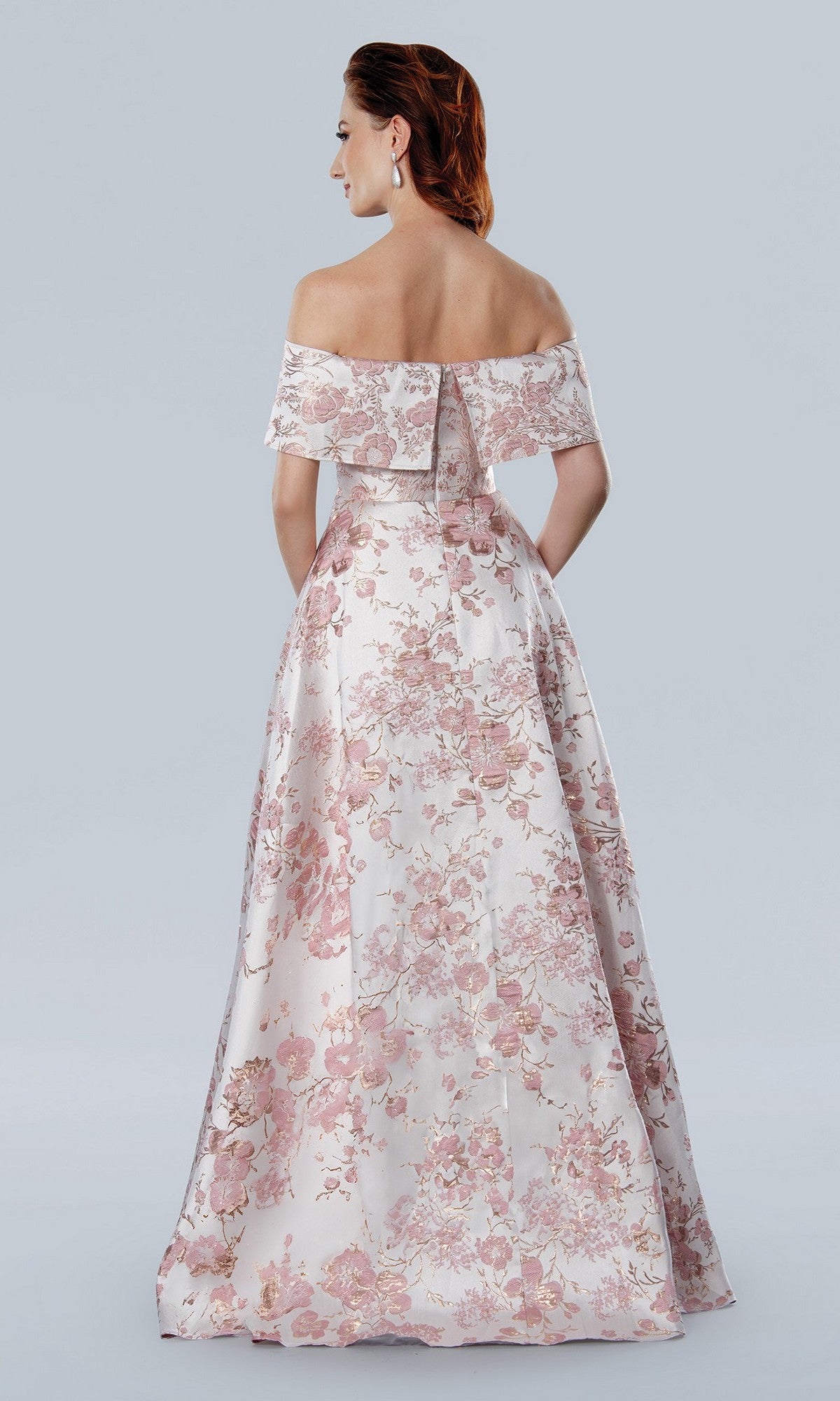 Woman wearing the Stella Couture 25303 long formal dress, a floor-length white gown with pink floral patterns and an off-the-shoulder neckline, stands with her back to the camera against a plain light background.